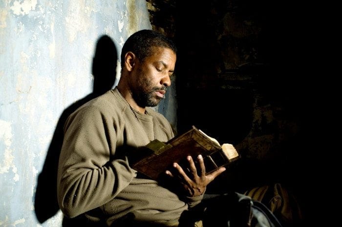 Eli (Denzel Washington) sitting against a wall reading during a scene in the Book of Eli