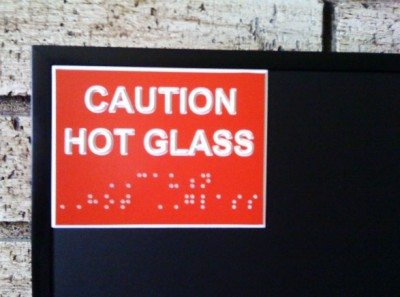 A sign that says "Caution Hot Glass" in print and braille