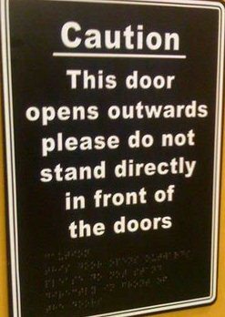 A print and braille sign that says "Caution. This door opens outwards, please do not stand directly in front of the doors."