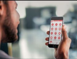 Individual smiling while holding up his smart phone that shows the destination options that the user can choose from such as hospitals, resturants, and transportation. 