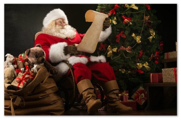 Santa sitting in a chair next to a Christmas tree reading a child's letter.