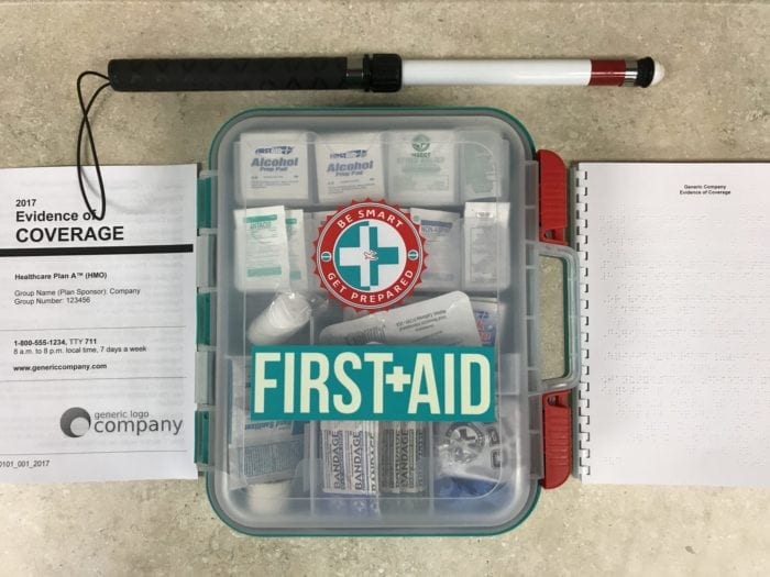 Braille and large print Evidence of Coverage documents on a counter top with a first aid kit and a white cane.