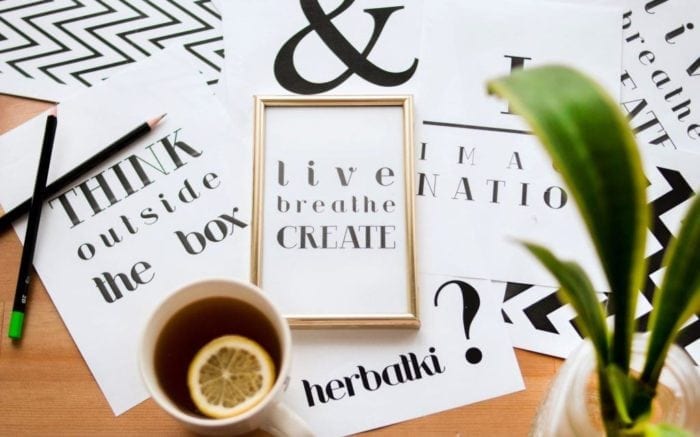 Table covered with paper, pencils, a mug of tea and a small plant. The papers read, "like breathe create" and "think outside the box"