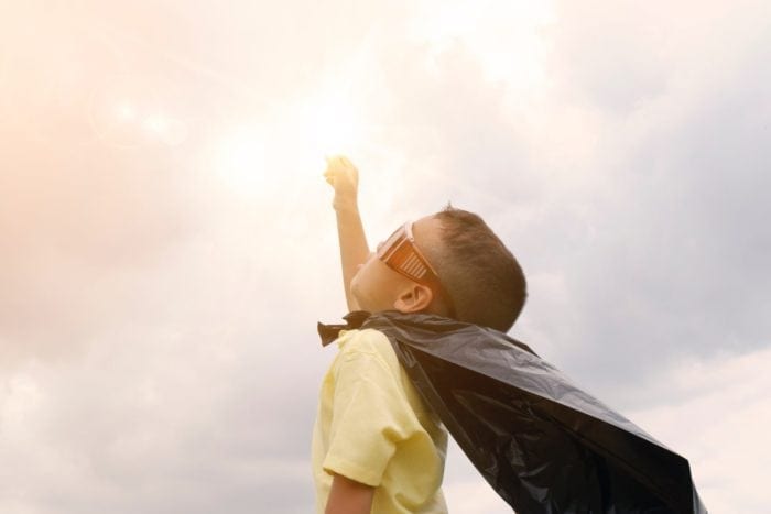 Young boy wearing a plastic bag cape and goggles holding his fist to a cloudy but bright sky