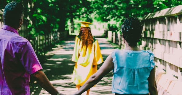 The back of a man and woman holding hands behind a female student in a cap and gown