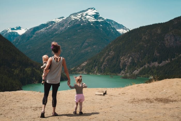 The backs of a woman holding a baby in one arm and a toddlers hand in the other overlooking a lake and snow-capped mountain