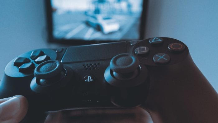 Close-up of a Playstation 4 controller with a blurred image of a video game on a TV in the background.