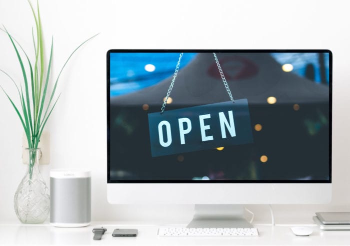 Computer monitor with an "Open" sign on the screen indicating an accessible website, keyboard, plant and other various desktop items.