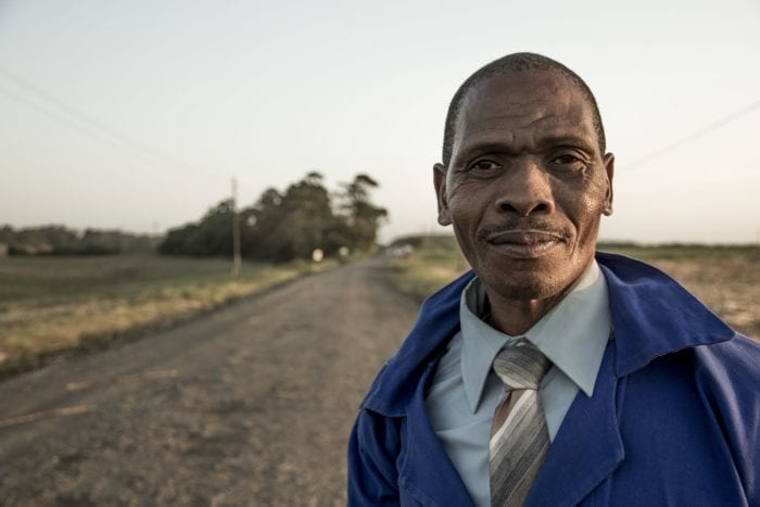 Elderly man looking ahead with an open road and field behind him