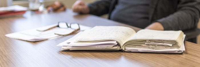 gentleman sitting at a desk with glasses on the table and a notebook full of papers.