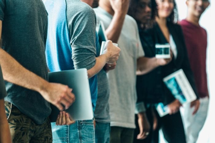 A group of casually dressed young adults stand in a row holding laptops and cups of coffee.