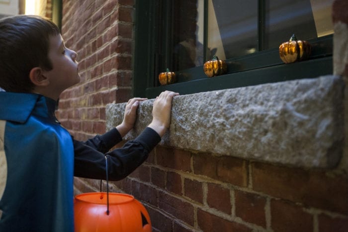 Young boy wearing a cape, holding a jack-o-lantern candy bucket, looking in a window with halloween decorations