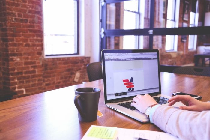 Person's arms and fingers typing on a laptop to add alt text to an image in an office warehouse and tea mug