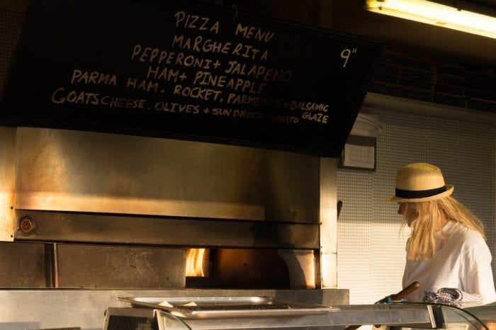 an employee at a pizza oven with the menu overhead written in chalk paint. 