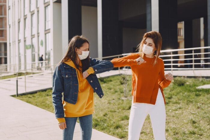 Two women outside wearing medical face masks and bumping elbows