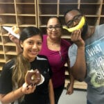 2 team members holding donuts and 1 team member holding a banana up to his mouth like a smile