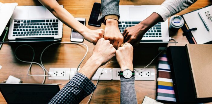 Five arms of colleagues joining fists over a table with laptops, chargers, notebooks to show teamwork and accomplishment.