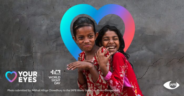 A girl with a big smile and eyes closed hugging another girl, who's also smiling, from behind. There's a heart around their heads and the World Sight Day, Love Your Eyes, and IAPB logos.