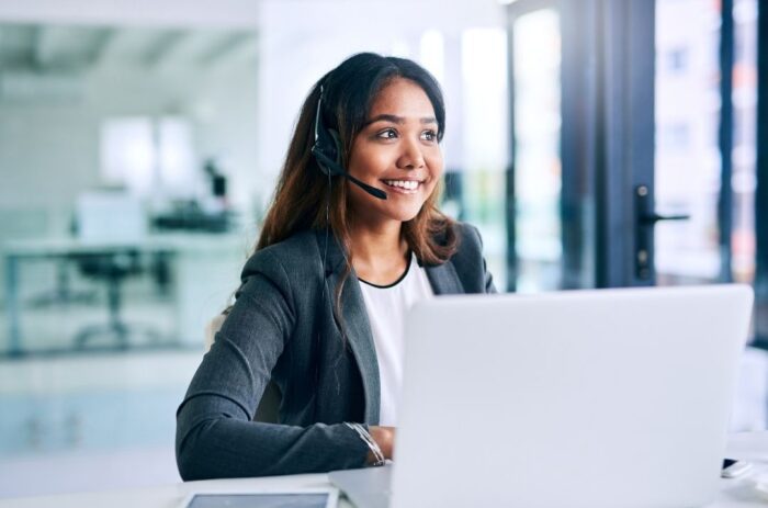 A woman with a headset on, typing on a laptop, sitting in an office.