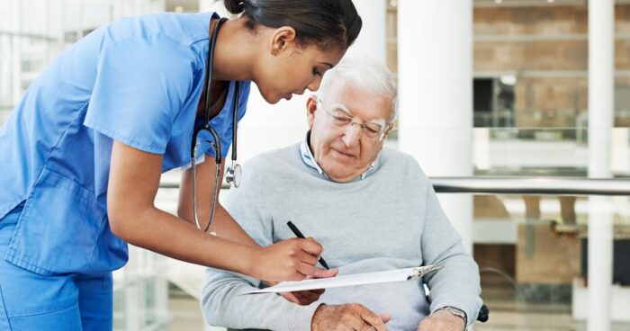 Nurse bending over and discussing paperwork that's on a clipboard with an elderly man in a wheelchair