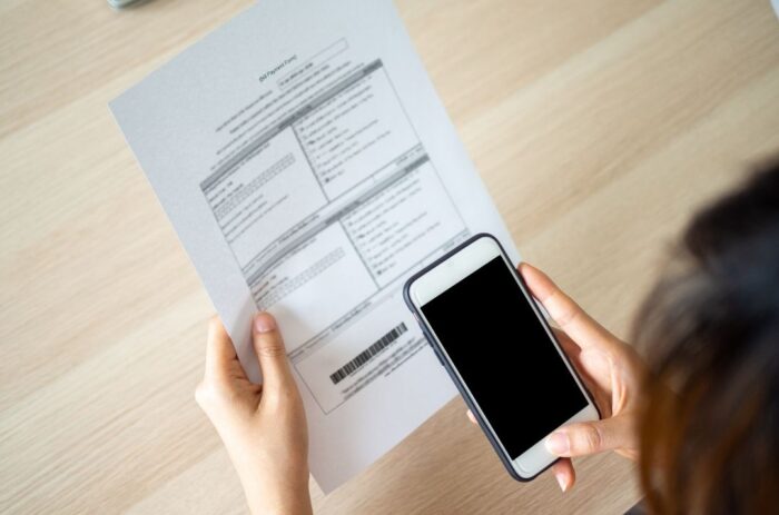 A person standing in front of a table holding a piece of paper with a barcode on it. The person is holding a phone, taking a picture of the barcode.